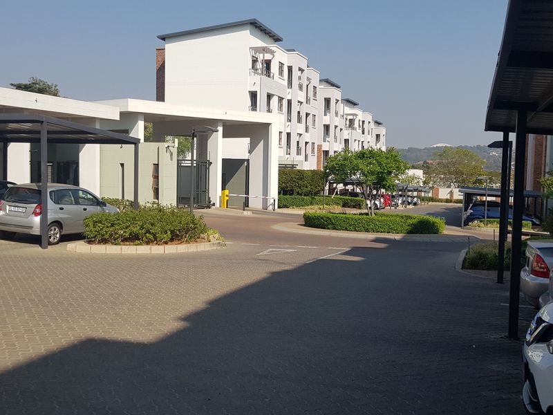 Exterior view of the apartment complex entrance featuring paved parking areas, covered carports, and well-maintained landscaped garden beds.