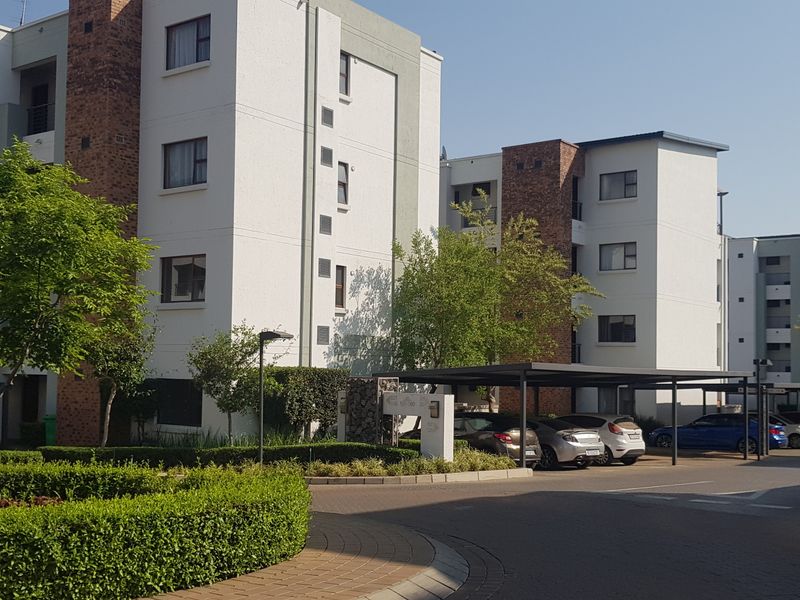 Exterior view of a modern apartment complex featuring multi-story residential blocks, paved driveways, covered parking bays, and well-maintained landscaped garden areas.