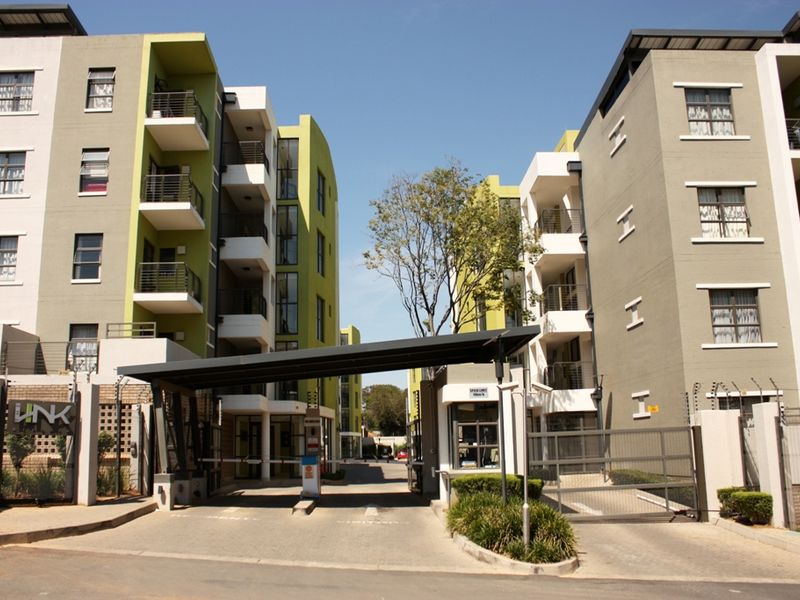 Front entrance view of a modern multi-story apartment complex featuring a secure gated access point, guard house, and paved driveway.