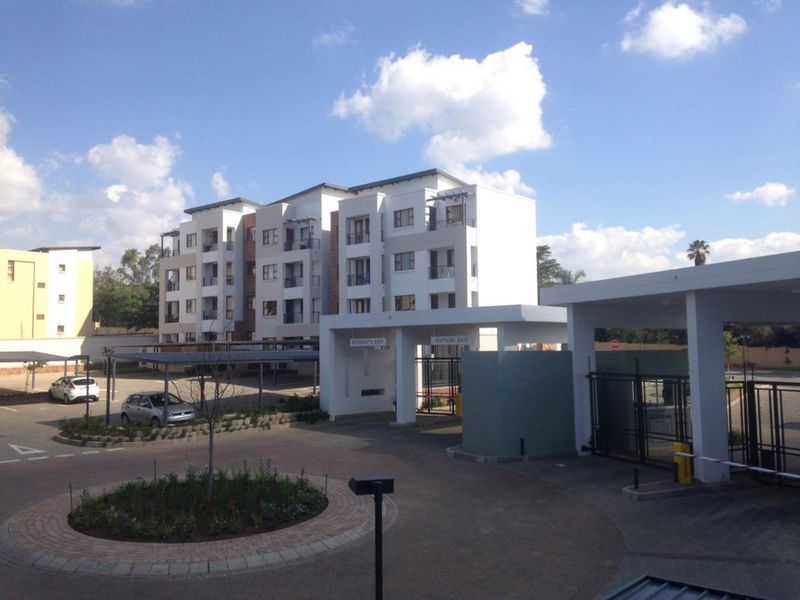 Exterior view of an apartment complex entrance featuring a secure boom gate, paved driveway, and covered parking bays.