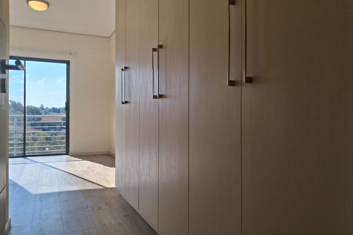 A bright, empty bedroom featuring modern built-in cupboards, laminate flooring, and a sliding glass door leading to a balcony with an outdoor view.