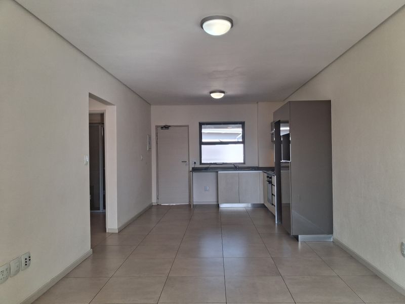 An open-plan living and kitchen area featuring neutral tiled flooring, built-in cabinetry, and integrated kitchen appliances in a clean, vacant space.