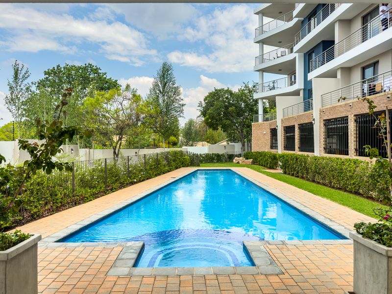 A well-maintained swimming pool area featuring brick paving, landscaped garden borders, and a view of the modern apartment building facade.