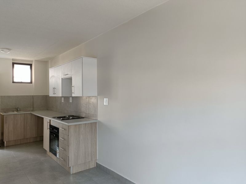 A clean, modern kitchen featuring light wood-finish cabinetry, a built-in oven and hob, tiled backsplash, and neutral floor tiles in an open-plan layout.