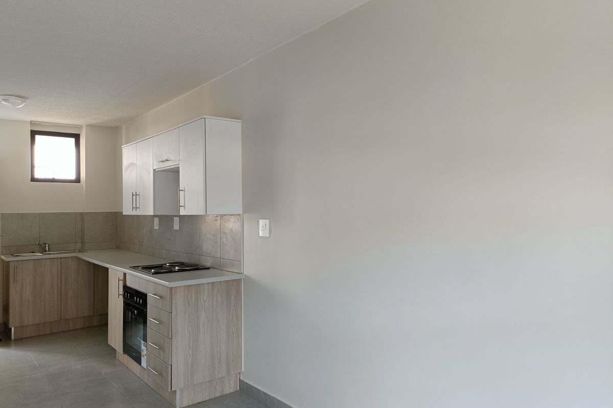 A clean, modern kitchen featuring light wood-finish cabinetry, a built-in oven and hob, tiled backsplash, and neutral floor tiles in an open-plan layout.