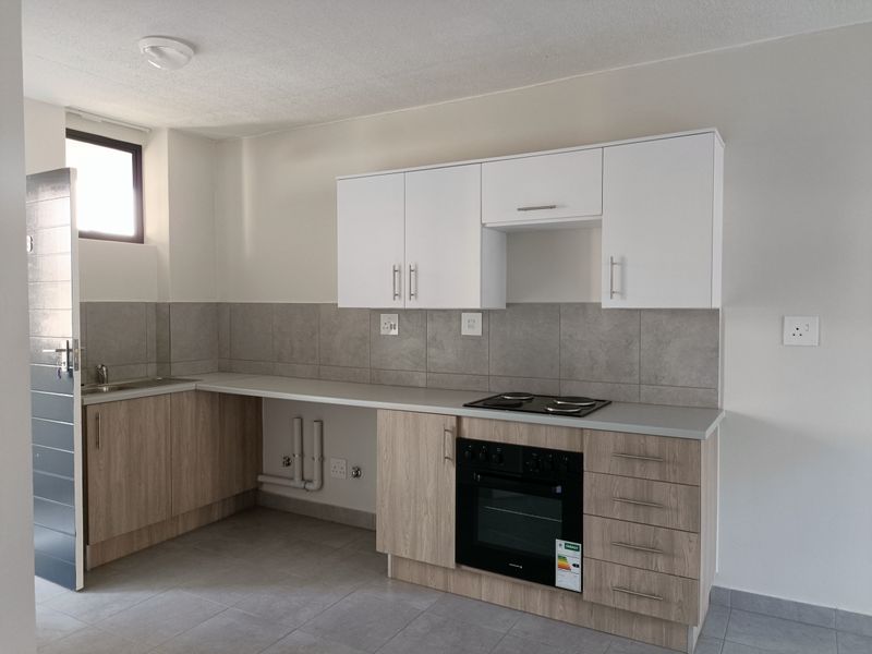 A clean, modern kitchen featuring light wood-finish cabinetry, white upper cabinets, grey tiled backsplash, and integrated electric oven and hob.