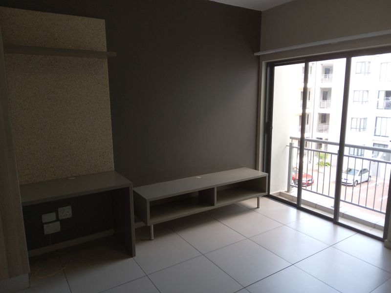 A bright living area featuring tiled flooring, a built-in media console, and a desk unit, with direct access to a balcony through sliding glass doors.