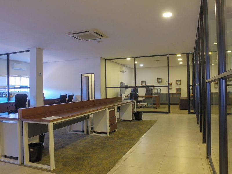 A modern open-plan office space featuring glass partitions, recessed lighting, integrated desk units, and a combination of tiled and carpeted flooring.