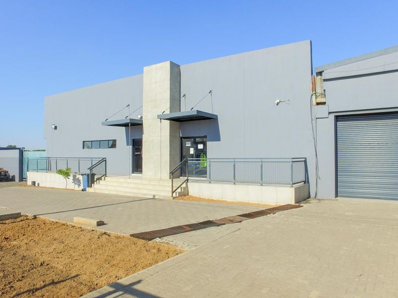 Modern industrial building facade featuring a large roller shutter door, paved driveway, and a raised entrance with metal railings and concrete stairs.