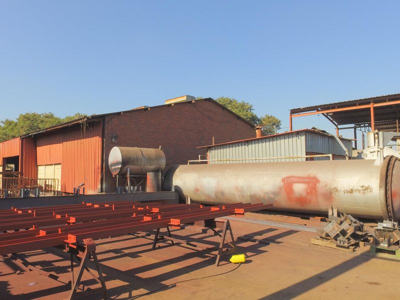 Industrial yard area featuring a large metal pressure vessel, structural steel beams, and brick-walled warehouse buildings with corrugated metal roofing.