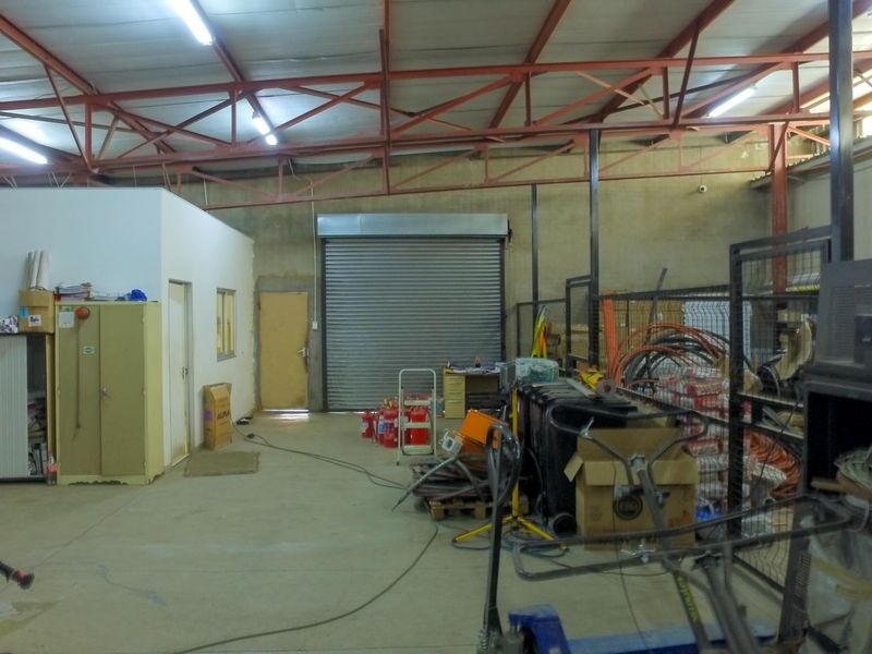 Industrial warehouse space featuring exposed steel roof trusses, concrete flooring, a roller shutter door, and an integrated internal office partition.