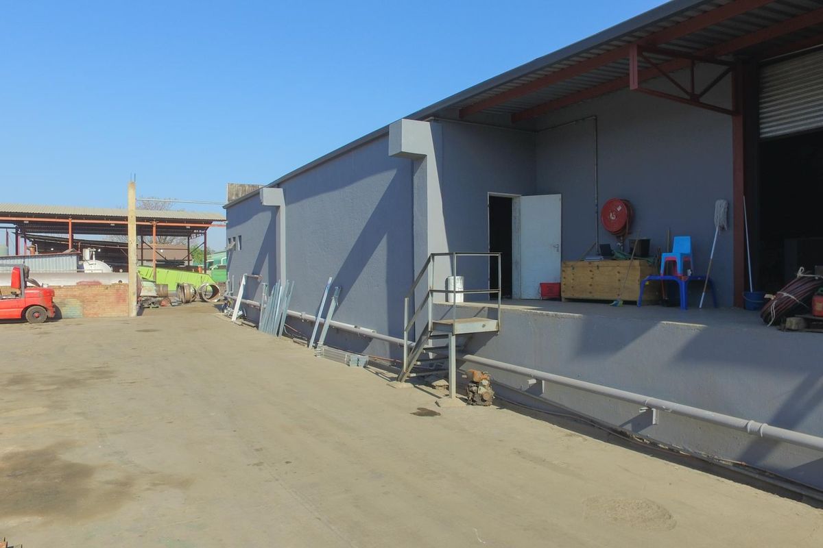 Industrial exterior view showing a loading dock area with a roller shutter door, steel staircase, and concrete yard space.