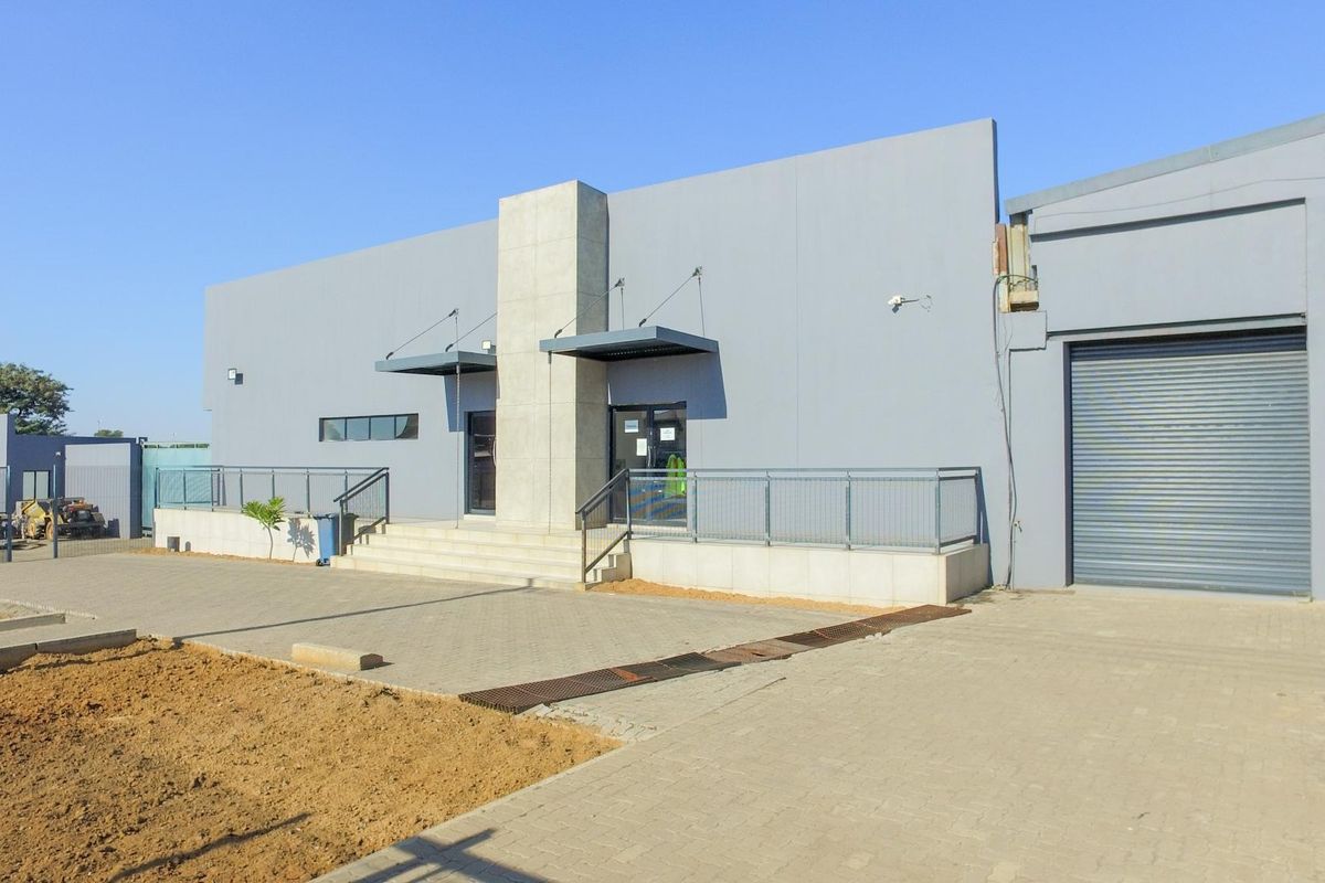 Modern industrial building facade featuring a large roller shutter door, paved driveway, and a raised entrance with metal railings and concrete stairs.