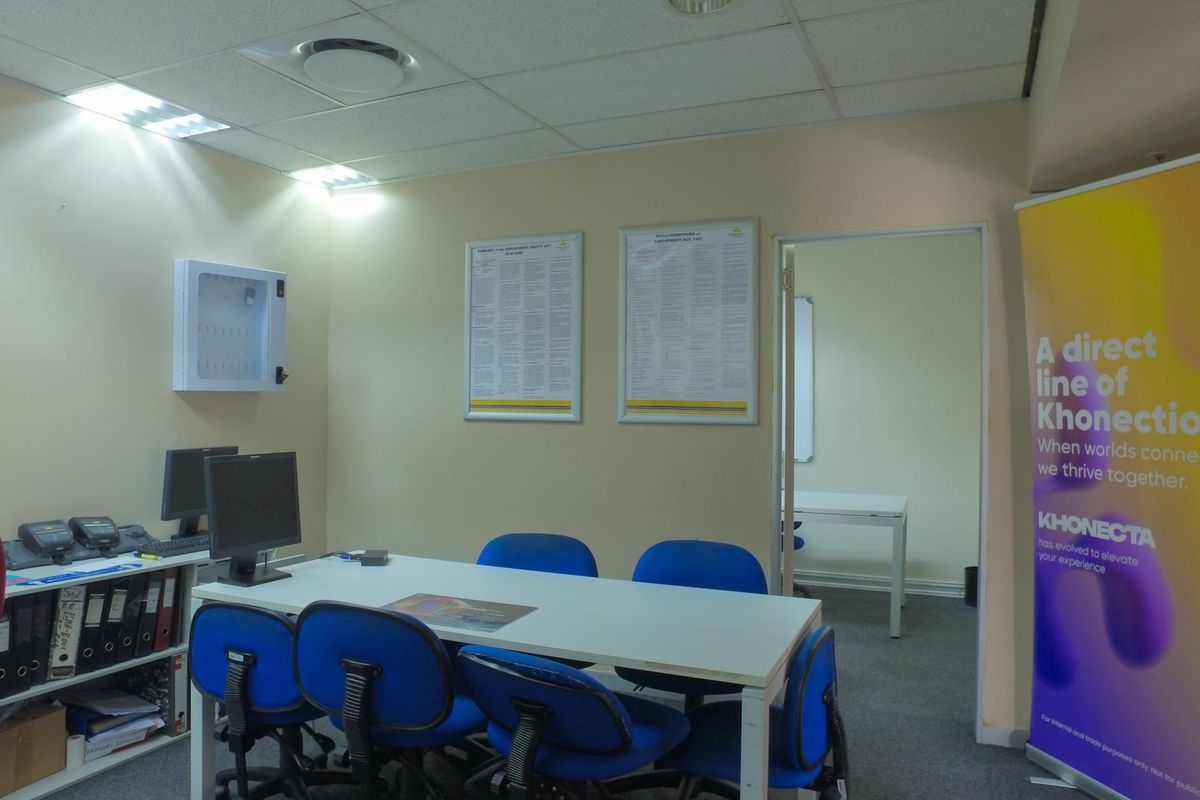 A functional office space featuring a suspended ceiling with recessed lighting, a wall-mounted key cabinet, and access to an adjoining room.