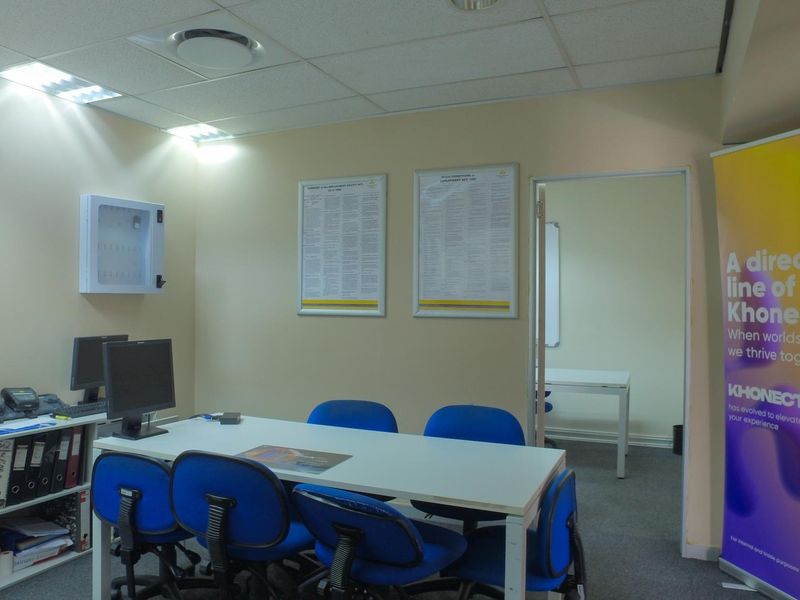A functional office space featuring a suspended ceiling with recessed lighting, a wall-mounted key cabinet, and access to an adjoining room.