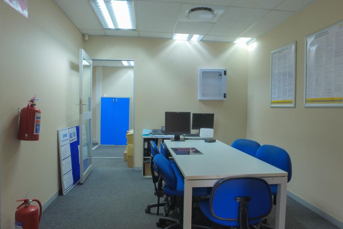 A functional office space featuring carpeted flooring, suspended ceiling with recessed lighting, and wall-mounted safety equipment.