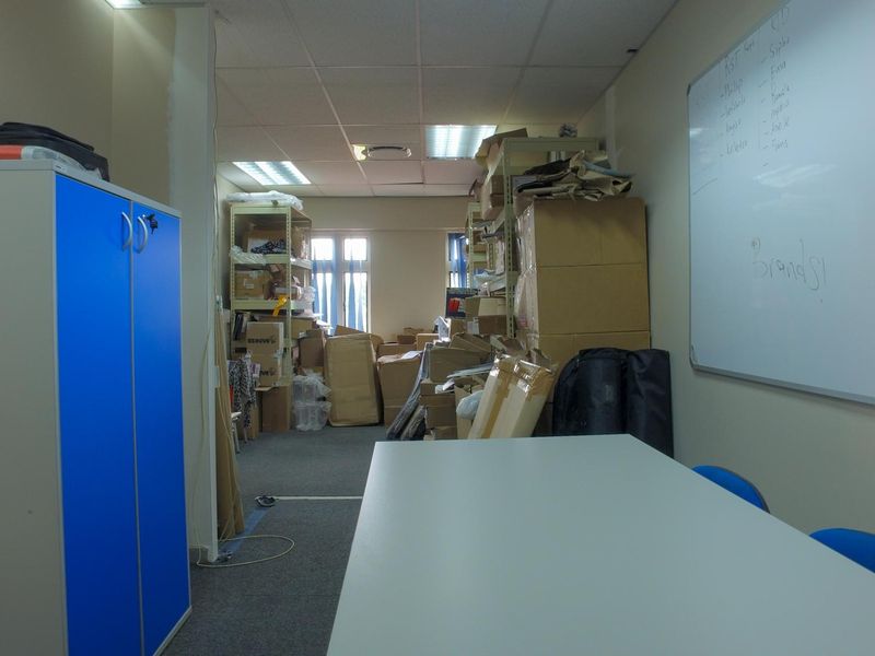 A functional office space featuring a suspended ceiling, recessed lighting, and a wall-mounted whiteboard, currently utilized for storage and workspace.