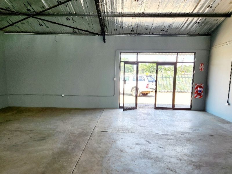 An open-plan office space featuring exposed steel roof trusses, polished concrete flooring, and large aluminium-framed glass doors providing natural light.