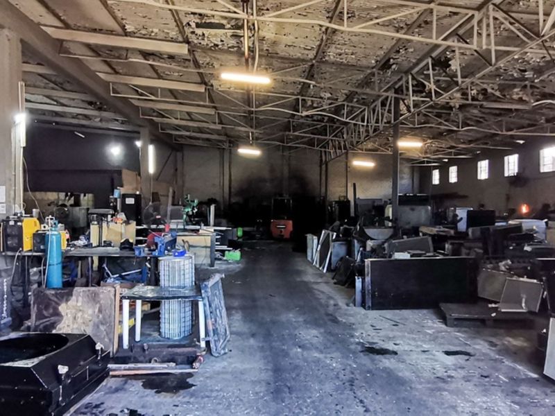 Large industrial warehouse space featuring exposed steel roof trusses, concrete flooring, and masonry walls, currently utilized for industrial operations.