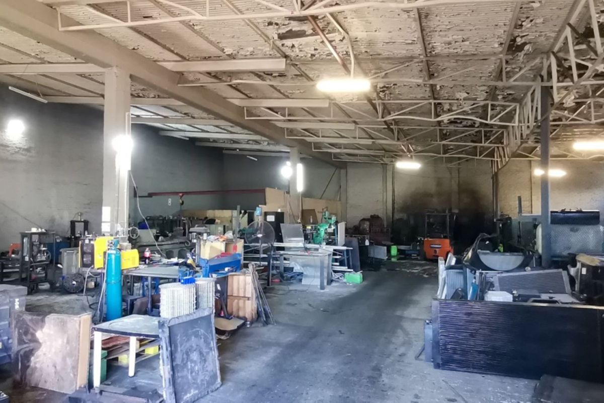 Industrial warehouse space featuring exposed steel roof trusses, concrete flooring, and masonry walls, currently utilized for workshop and storage purposes.