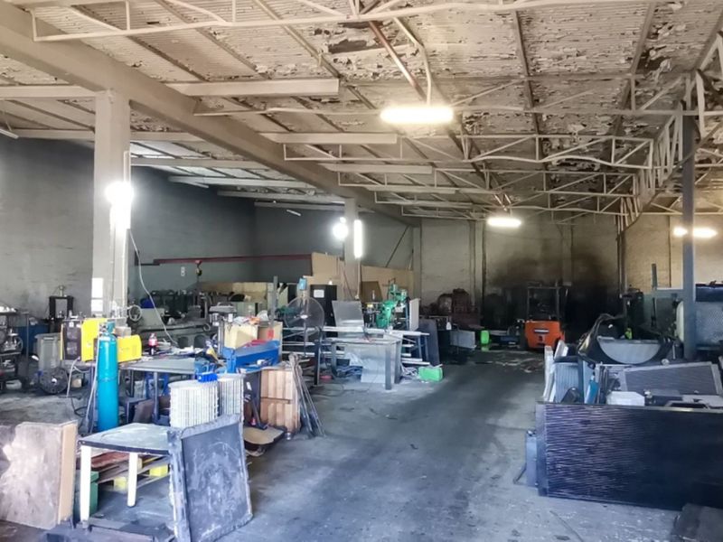 Industrial warehouse space featuring exposed steel roof trusses, concrete flooring, and masonry walls, currently utilized for workshop and storage purposes.