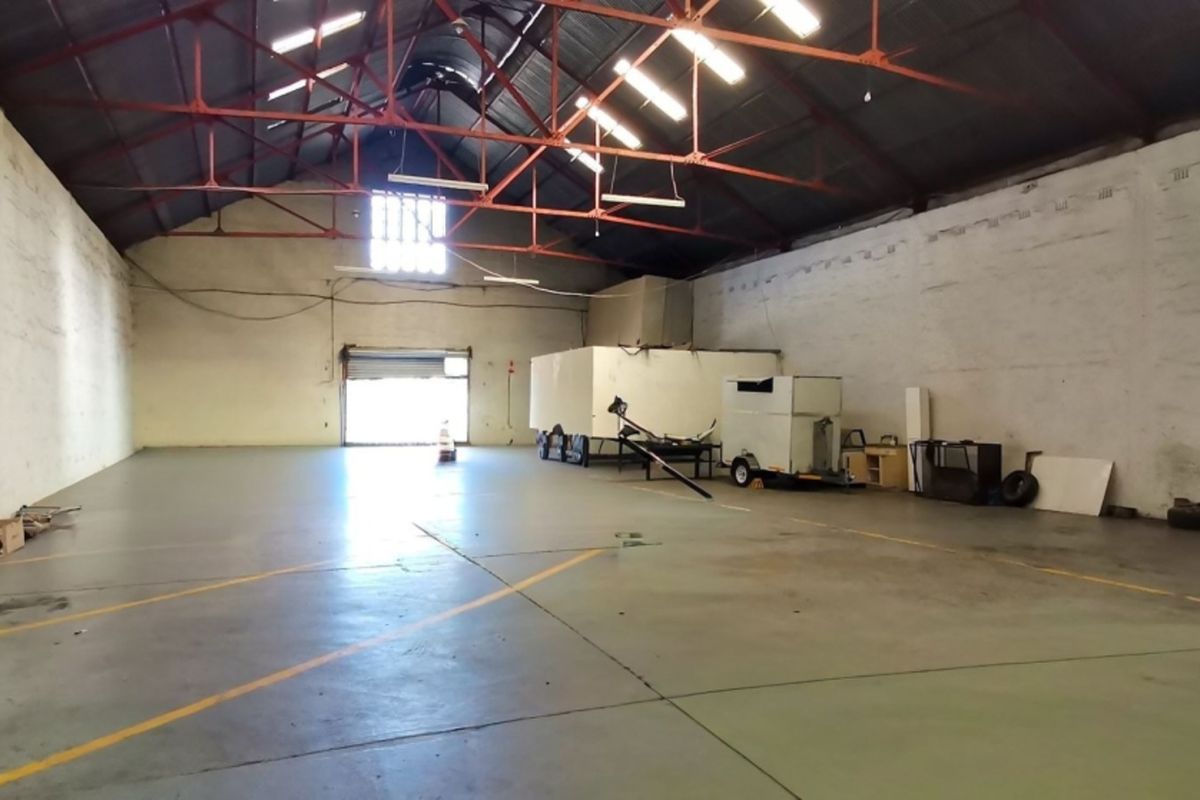 Spacious industrial warehouse unit featuring high ceilings with exposed steel trusses, concrete flooring, and a large roller shutter door for vehicle access.