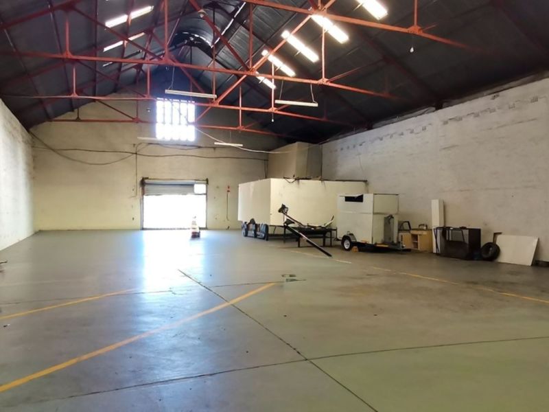 Spacious industrial warehouse unit featuring high ceilings with exposed steel trusses, concrete flooring, and a large roller shutter door for vehicle access.