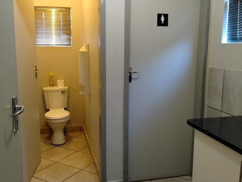 A functional office restroom featuring a toilet, vanity with basin, tiled flooring, and window blinds.
