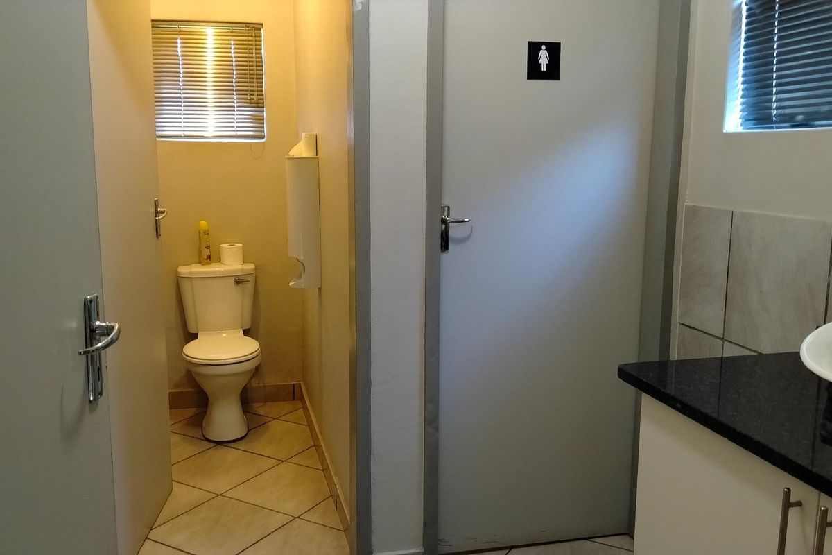 A functional office restroom featuring a toilet, vanity with basin, tiled flooring, and window blinds.