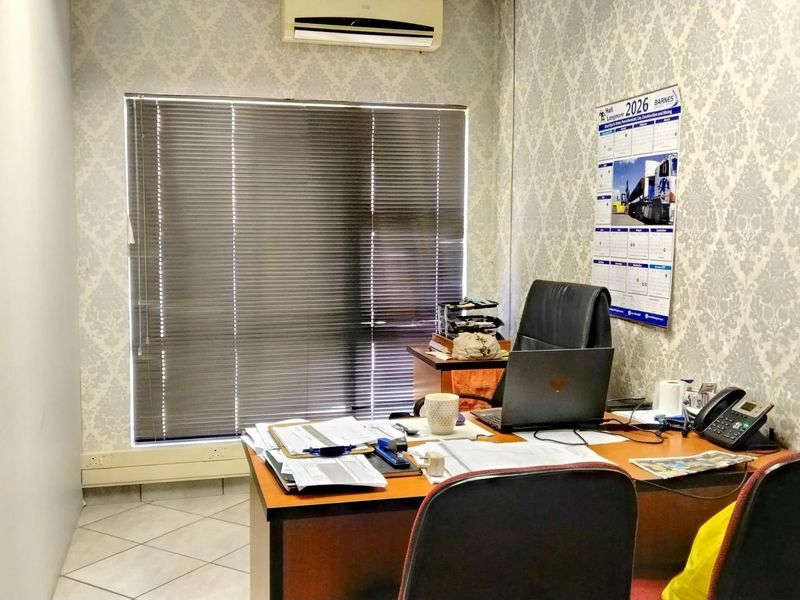 A functional office space featuring tiled flooring, a wall-mounted air conditioning unit, and large windows fitted with venetian blinds.