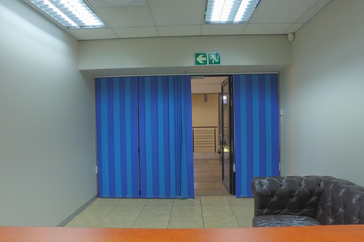 An office space featuring tiled flooring, recessed ceiling lighting, and blue vertical blinds leading to an adjacent corridor.