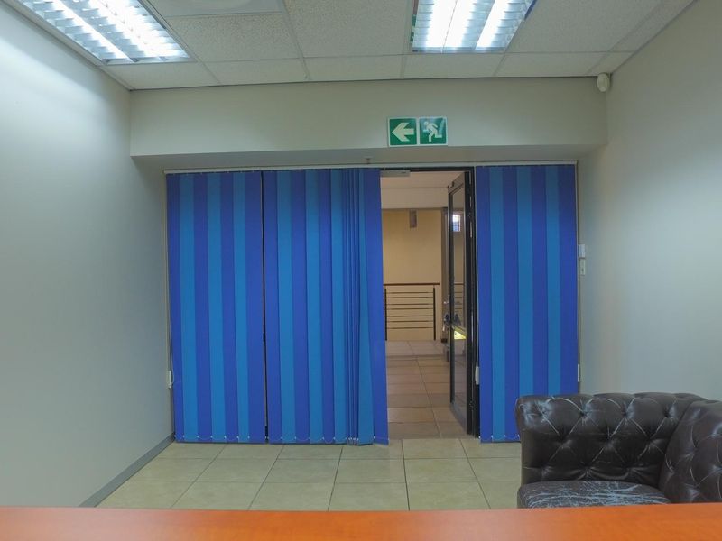An office space featuring tiled flooring, recessed ceiling lighting, and blue vertical blinds leading to an adjacent corridor.