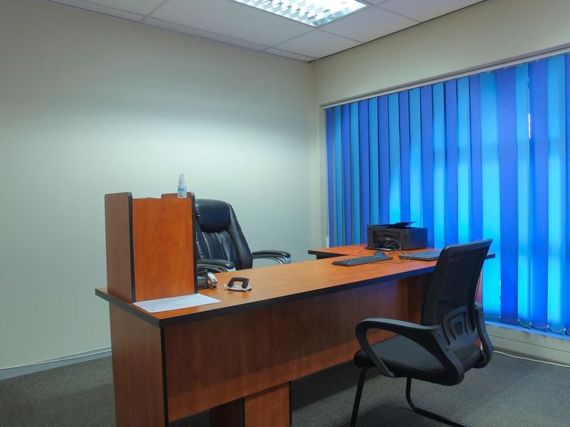 A functional office space featuring carpeted flooring, a suspended ceiling with recessed lighting, and large windows fitted with vertical blinds.