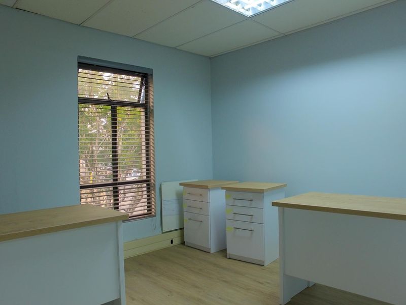 A functional office space featuring laminate flooring, a suspended ceiling with recessed lighting, and a window fitted with venetian blinds.