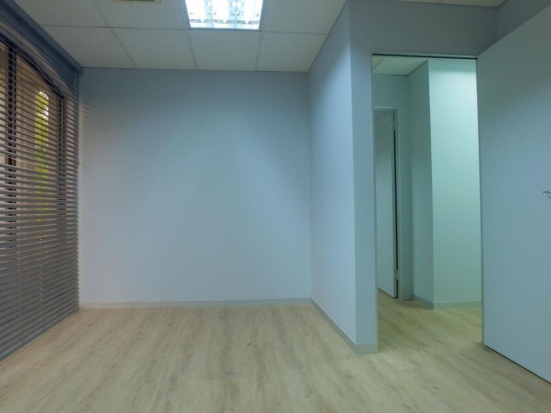 A clean, empty office space featuring light-toned laminate flooring, a suspended ceiling with recessed lighting, and venetian blinds on the window.