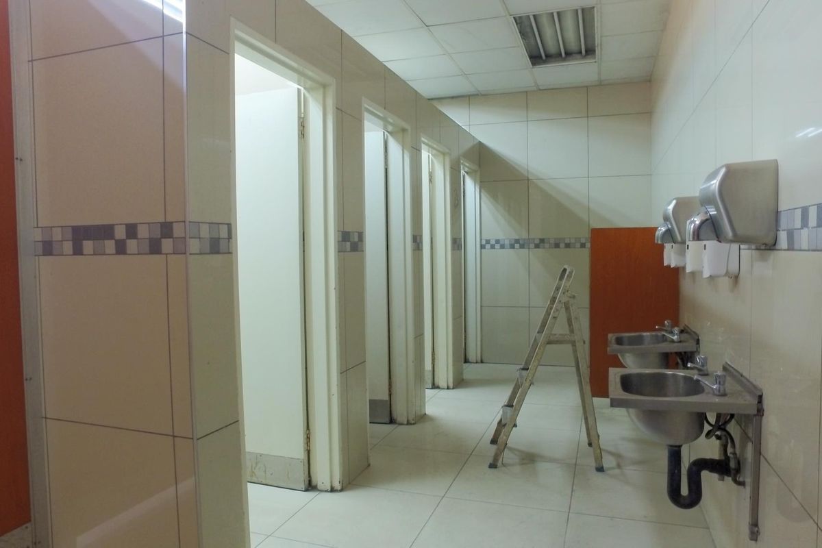 A functional industrial-style communal bathroom featuring multiple cubicles, stainless steel wall-mounted sinks, and tiled finishes throughout.