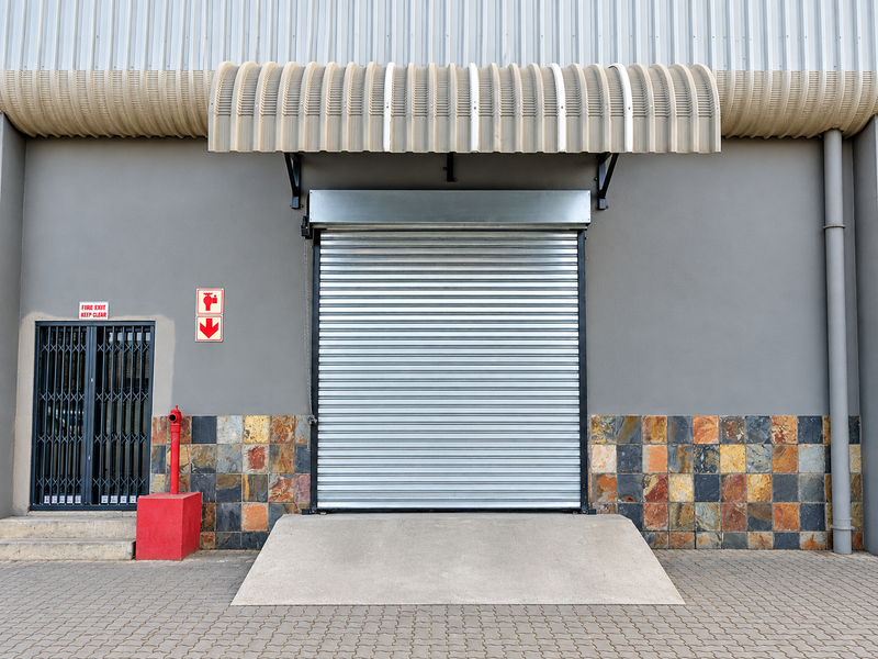 Industrial rear exterior featuring a large roller shutter door, concrete loading ramp, security gate, and fire hydrant access against a stone-clad facade.