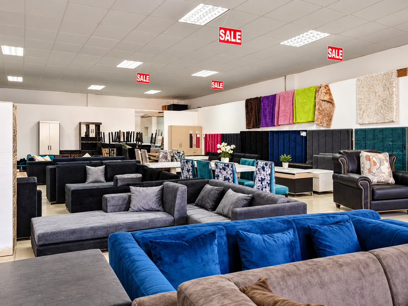 A spacious retail showroom featuring a suspended ceiling grid with recessed lighting and neutral tiled flooring, currently set up as a furniture display area.