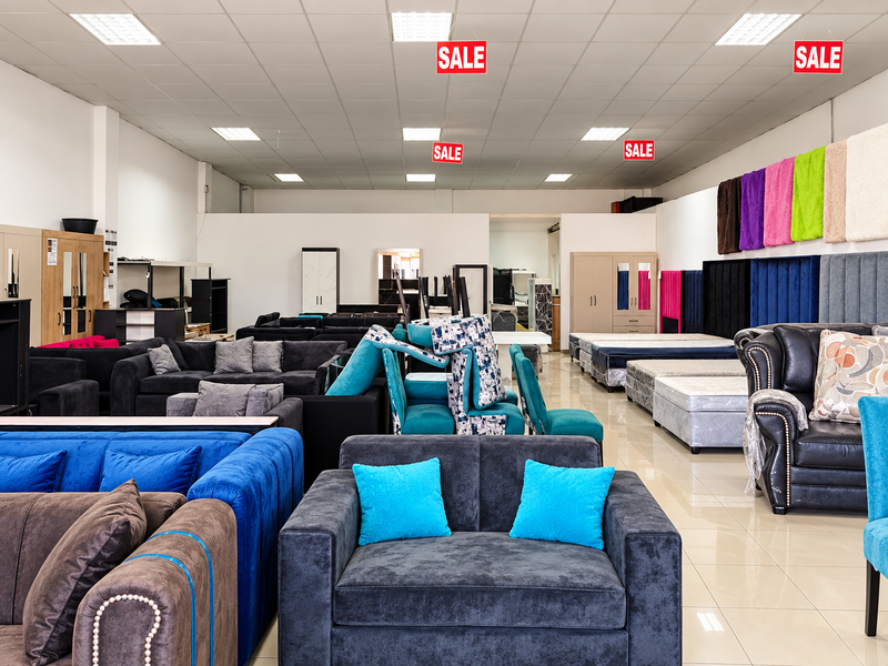 A spacious retail showroom featuring a suspended ceiling with recessed lighting and polished tiled flooring, currently utilized for furniture display.