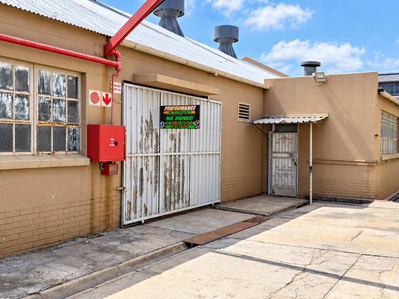 Rear industrial exterior featuring a large sliding access door, fire safety equipment, and concrete yard space with visible wear to finishes.