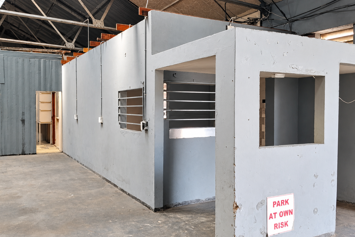 An industrial utility space featuring exposed steel roof trusses, concrete flooring, and partitioned office or security structures with surface-mounted electrical conduits.