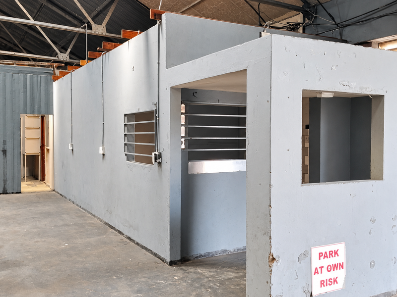 An industrial utility space featuring exposed steel roof trusses, concrete flooring, and partitioned office or security structures with surface-mounted electrical conduits.