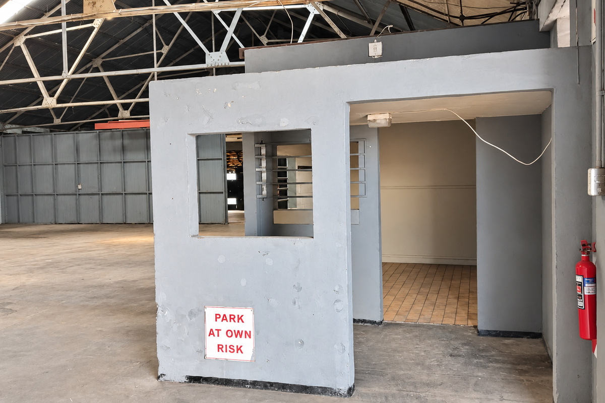 Industrial warehouse interior featuring a central security booth, exposed steel roof trusses, and a combination of concrete and tiled flooring.