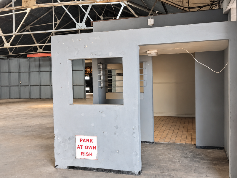 Industrial warehouse interior featuring a central security booth, exposed steel roof trusses, and a combination of concrete and tiled flooring.
