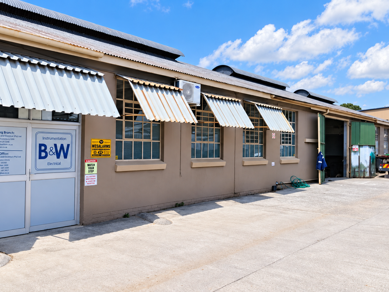 Industrial facility exterior featuring concrete driveway, steel-framed windows with corrugated metal awnings, and secure access points.