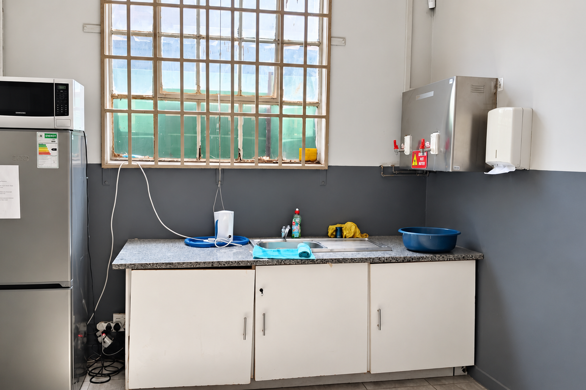 A functional industrial kitchenette featuring built-in cabinetry, a stainless steel sink, granite countertop, and a wall-mounted water dispenser with security bars on the window.
