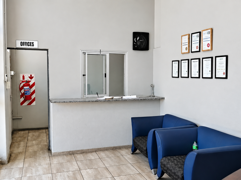 A functional industrial reception area featuring a service counter, tiled flooring, and a dedicated office access point with safety equipment.