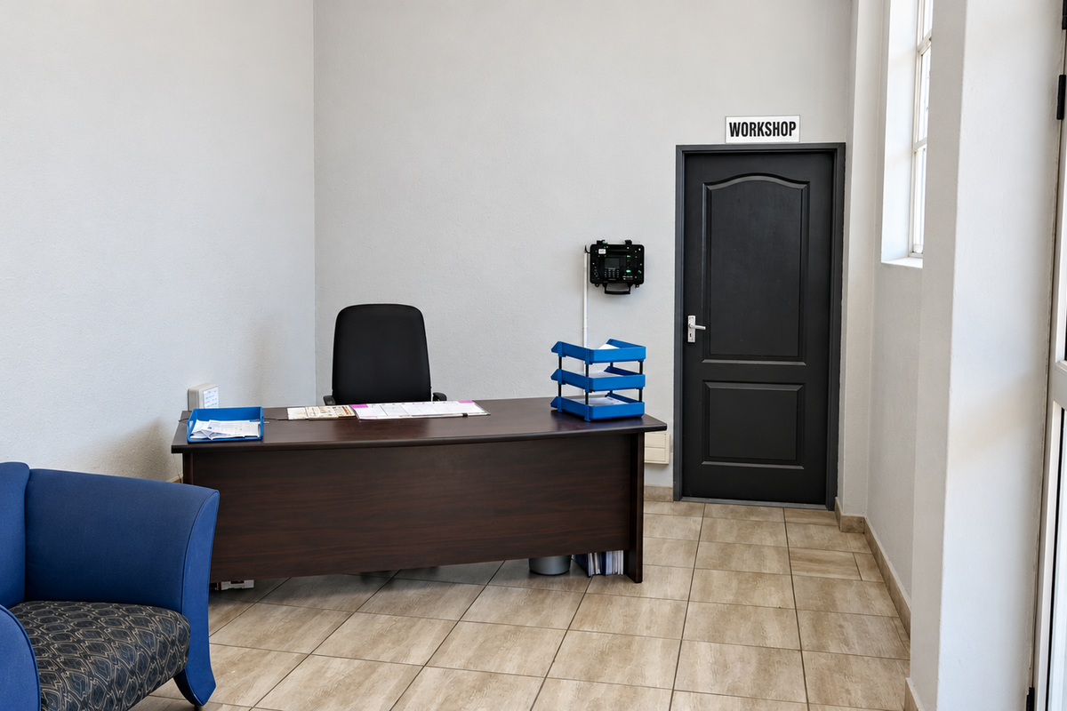 A functional office space featuring tiled flooring, a secure internal door leading to the workshop, and natural light from the adjacent window and entrance.