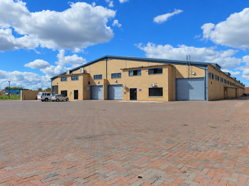 Industrial warehouse facility featuring a large paved yard, multiple roller shutter doors, brick exterior, and secure perimeter fencing.