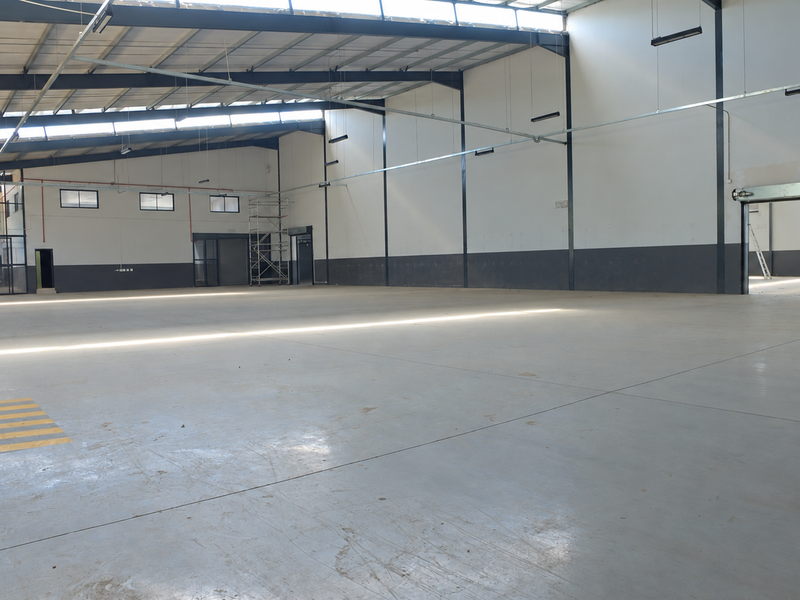 Spacious industrial warehouse featuring high ceilings with exposed steel trusses, polished concrete flooring, and multiple access points via roller shutter doors.